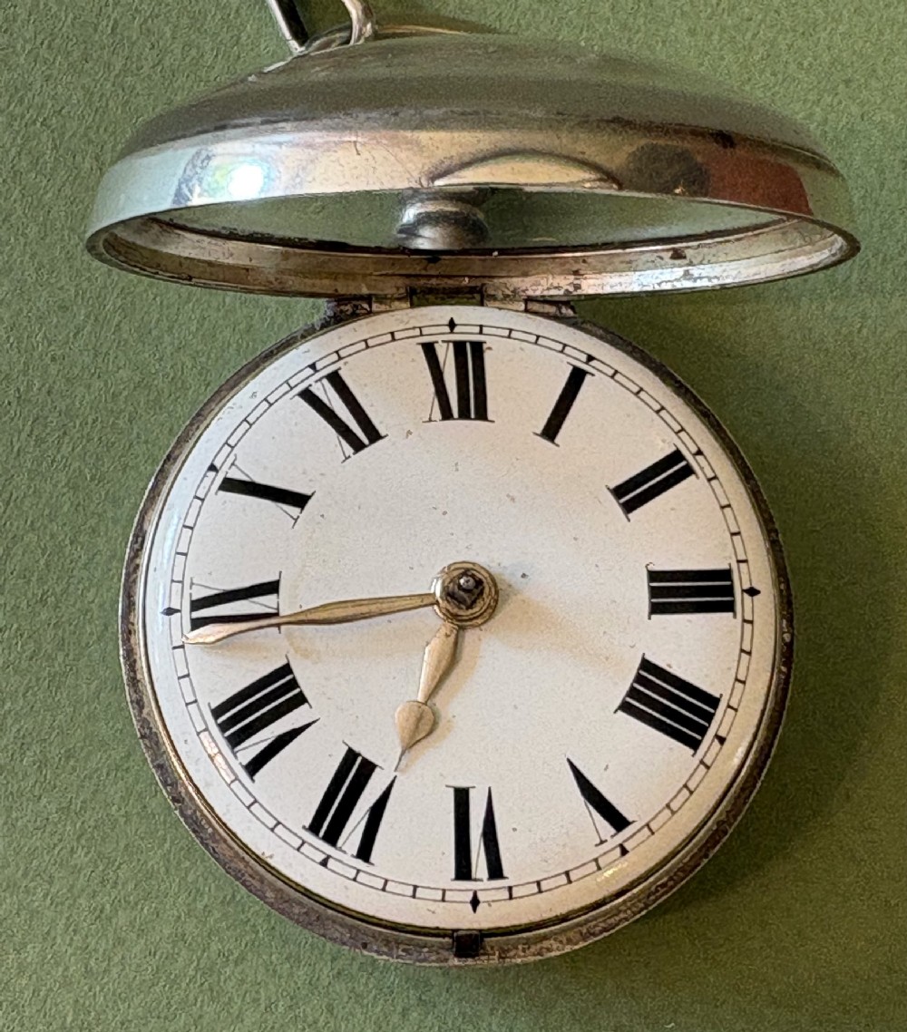 An antique silver pair-cased pocket watch featuring a white enamel dial with black Roman numerals, connected to a silver chain formed of linked coins, a T-bar, and a small cross charm. View III