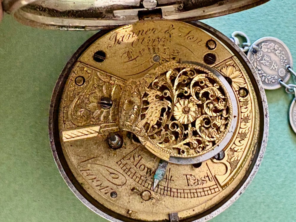 An antique silver pair-cased pocket watch featuring a white enamel dial with black Roman numerals, connected to a silver chain formed of linked coins, a T-bar, and a small cross charm. View IV