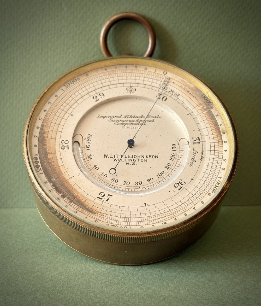 An antique circular brass pocket aneroid barometer and altimeter with a white engraved dial and suspension ring top. View II