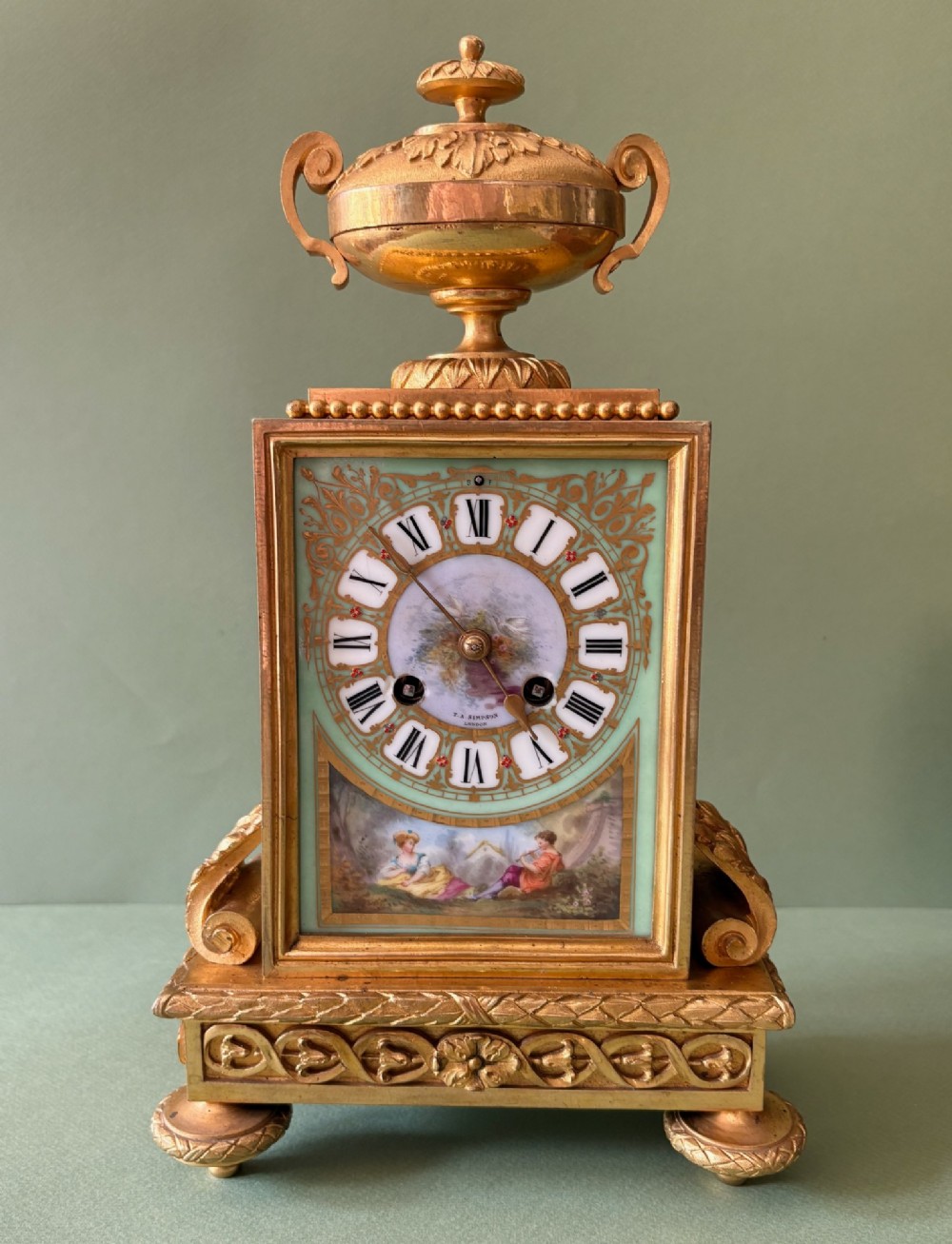 A 19th-century French gilt metal mantel clock featuring hand-painted green porcelain panels and an urn finial, displayed inside a glass dome on a wooden base. View II
