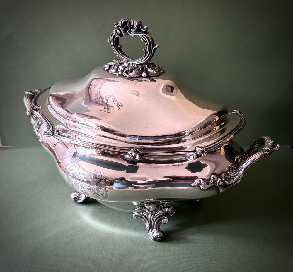An antique silver-plated three-piece soup tureen featuring a domed lid with a scroll handle, engraved heraldic crest, and ornate scrolled feet. View VI