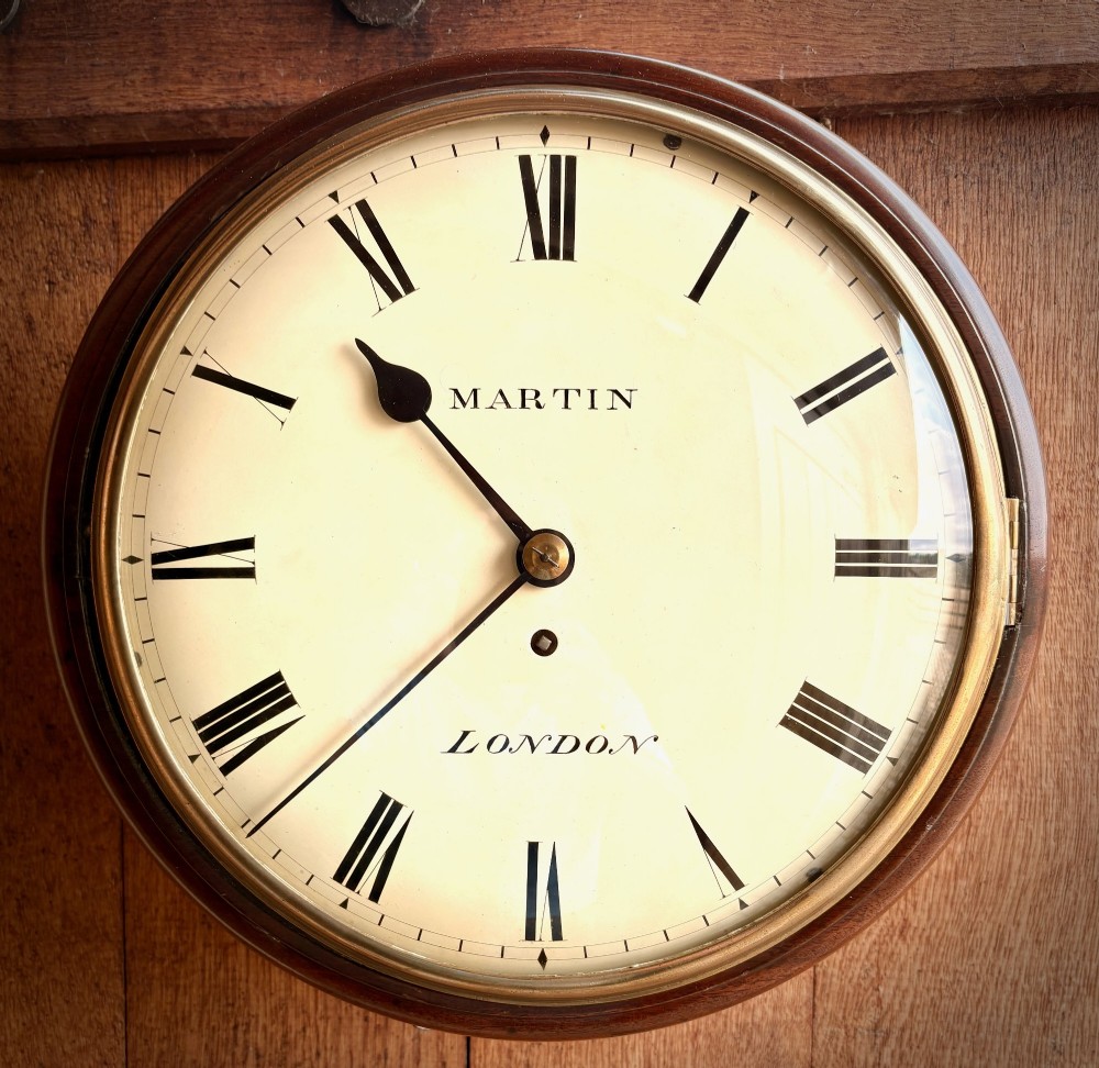 A circular Regency period mahogany wall clock featuring a brass bezel, off-white dial, black Roman numerals, and black steel hands.