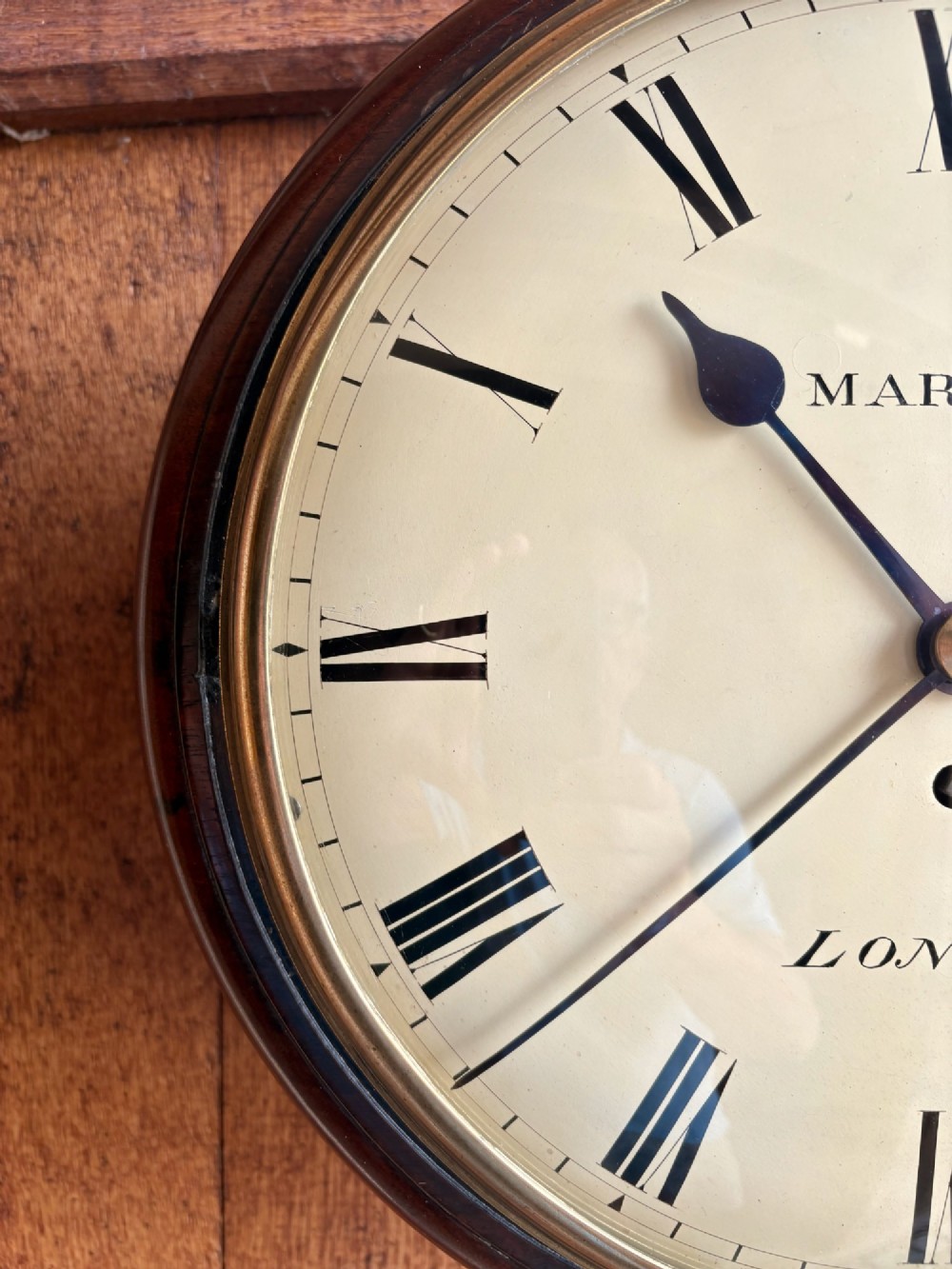 A circular Regency period mahogany wall clock featuring a brass bezel, off-white dial, black Roman numerals, and black steel hands. View II