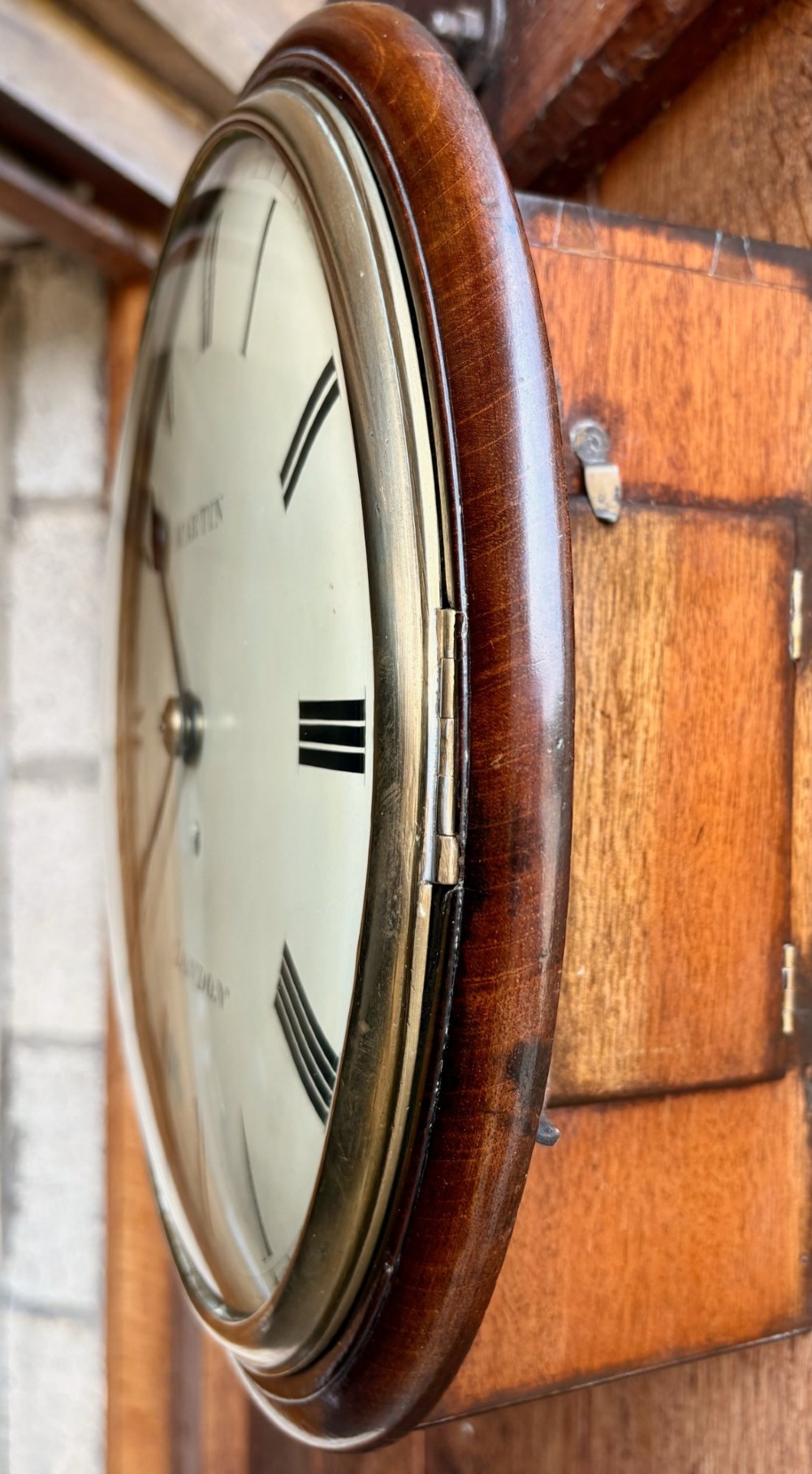 A circular Regency period mahogany wall clock featuring a brass bezel, off-white dial, black Roman numerals, and black steel hands. View III