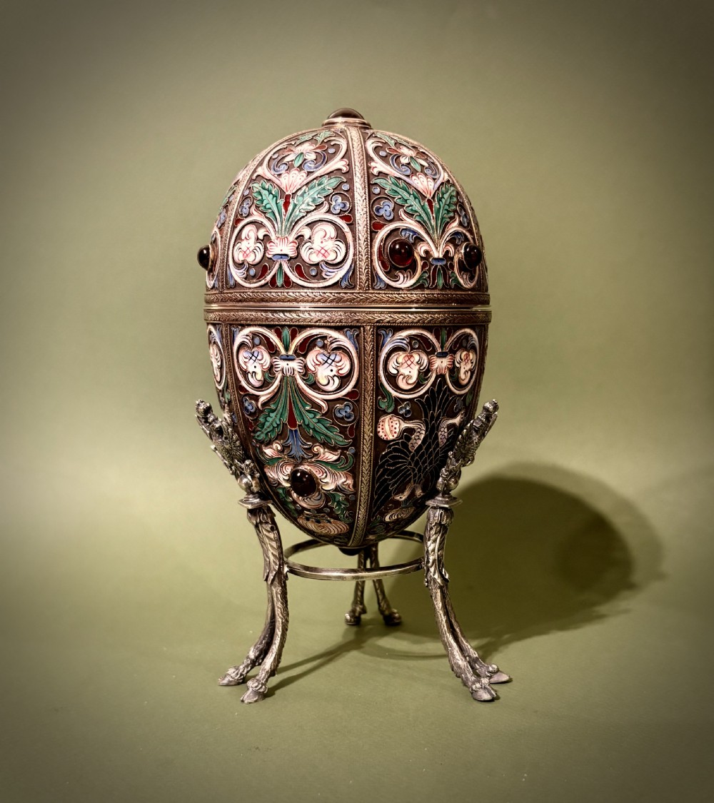 A Russian silver egg decorated with polychrome floral enamel scrolls and set with cabochon stones, resting on a tripod silver stand with hoofed feet.