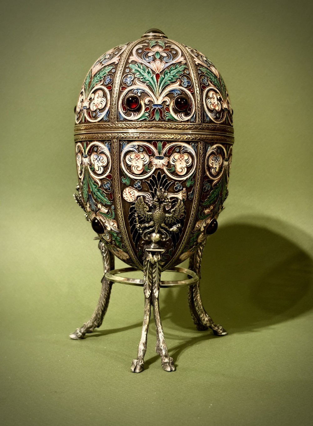A Russian silver egg decorated with polychrome floral enamel scrolls and set with cabochon stones, resting on a tripod silver stand with hoofed feet. View II
