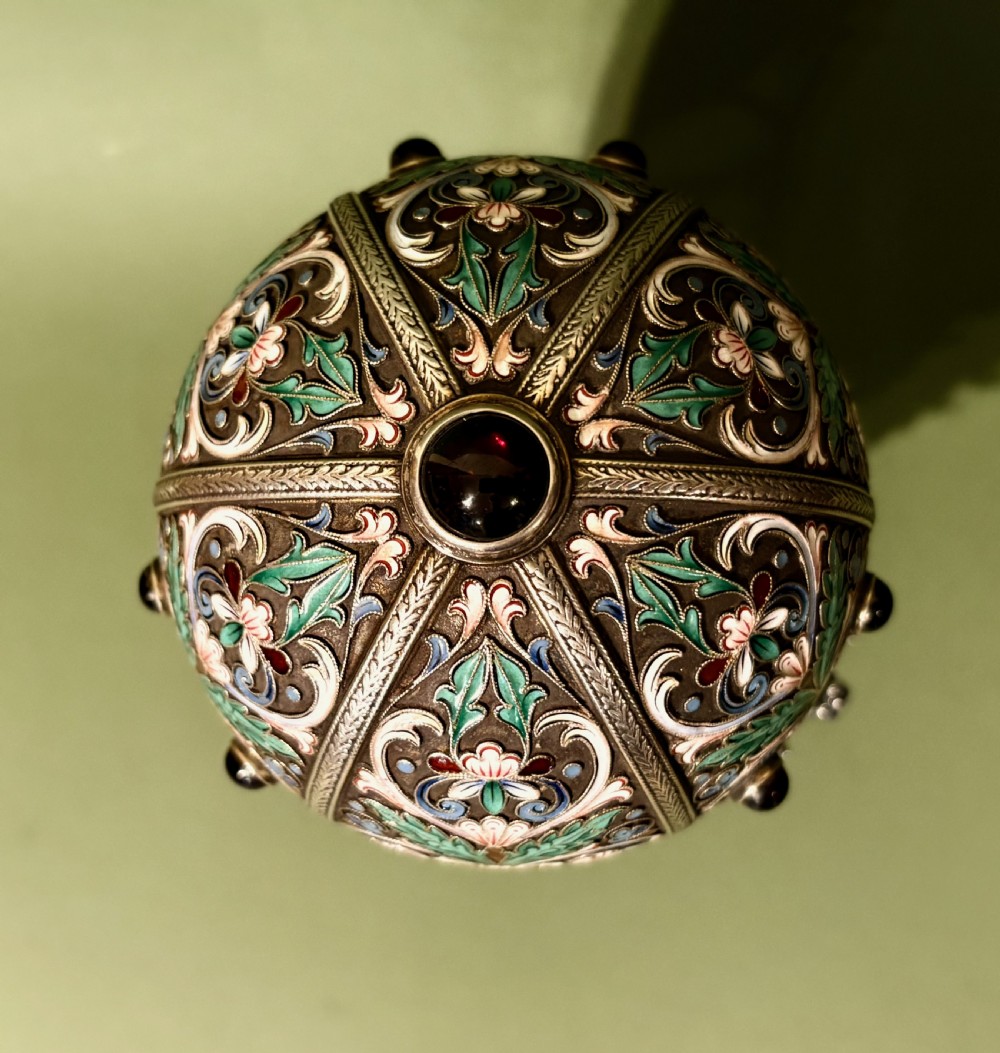A Russian silver egg decorated with polychrome floral enamel scrolls and set with cabochon stones, resting on a tripod silver stand with hoofed feet. View V