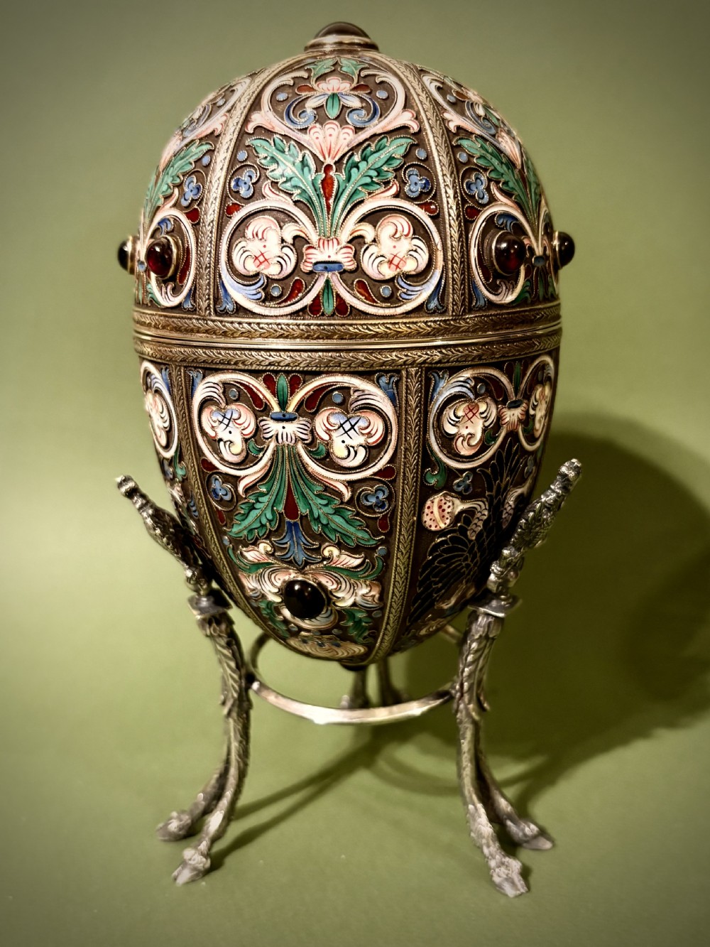 A Russian silver egg decorated with polychrome floral enamel scrolls and set with cabochon stones, resting on a tripod silver stand with hoofed feet. View XII