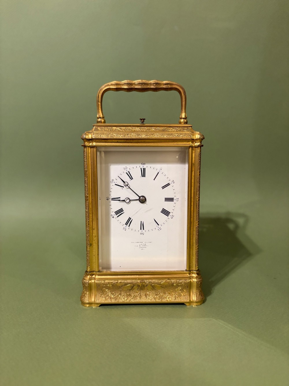 An antique rectangular French carriage clock featuring a gilt brass case with engraved foliage, glass side panels, a top carrying handle, and a white enamel dial displaying Roman numerals.