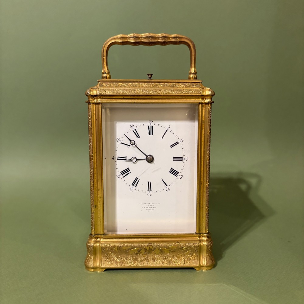 An antique rectangular French carriage clock featuring a gilt brass case with engraved foliage, glass side panels, a top carrying handle, and a white enamel dial displaying Roman numerals. View IX