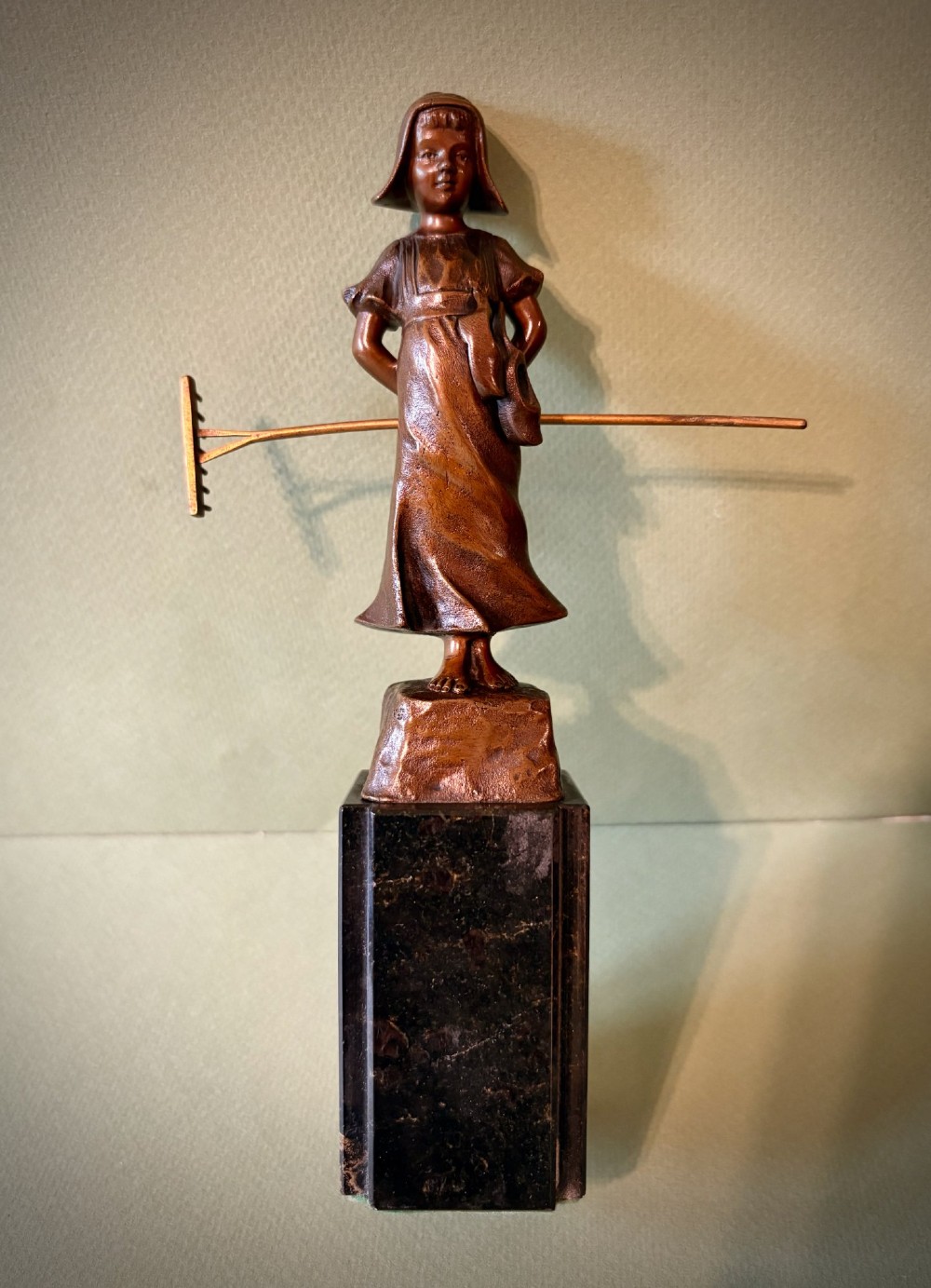 A patinated bronze figurine depicting a young farmer girl holding a rake, mounted on a dark marble pedestal base.