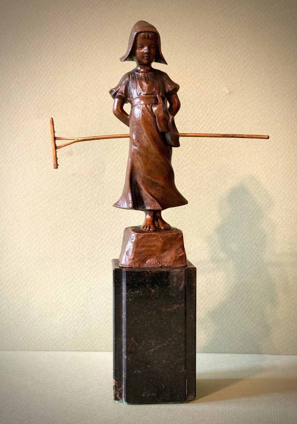 A patinated bronze figurine depicting a young farmer girl holding a rake, mounted on a dark marble pedestal base. View III