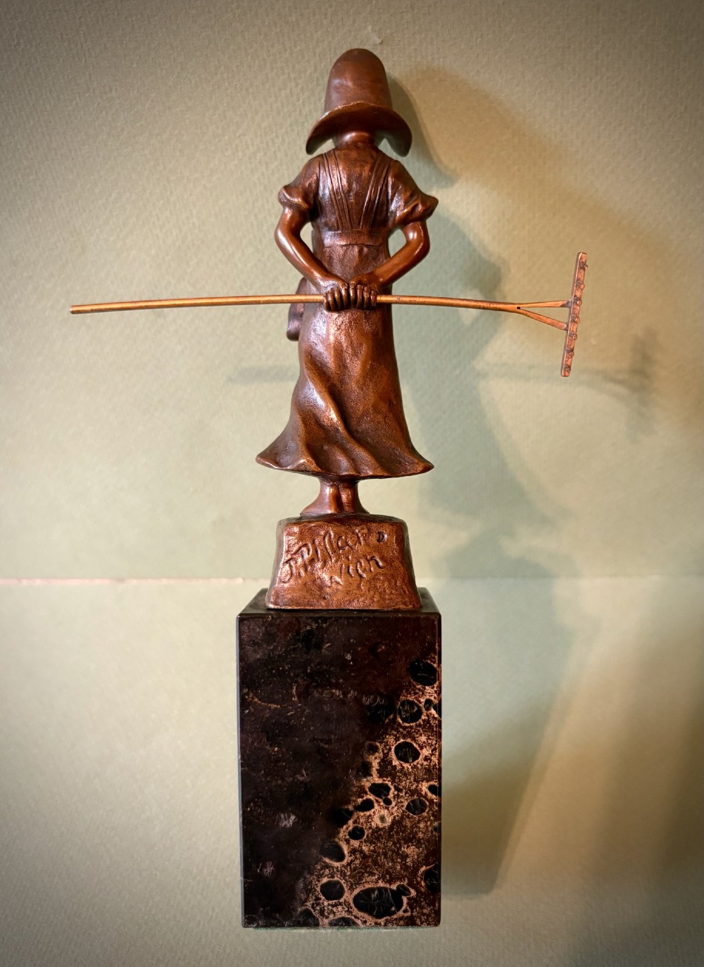 A patinated bronze figurine depicting a young farmer girl holding a rake, mounted on a dark marble pedestal base. View IV