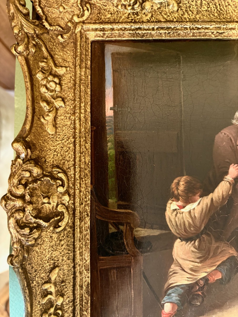 A nineteenth-century narrative oil painting on a wooden panel depicting a grandfather visiting a mother and children in a cottage interior, framed in an ornate gold gilt frame. View V