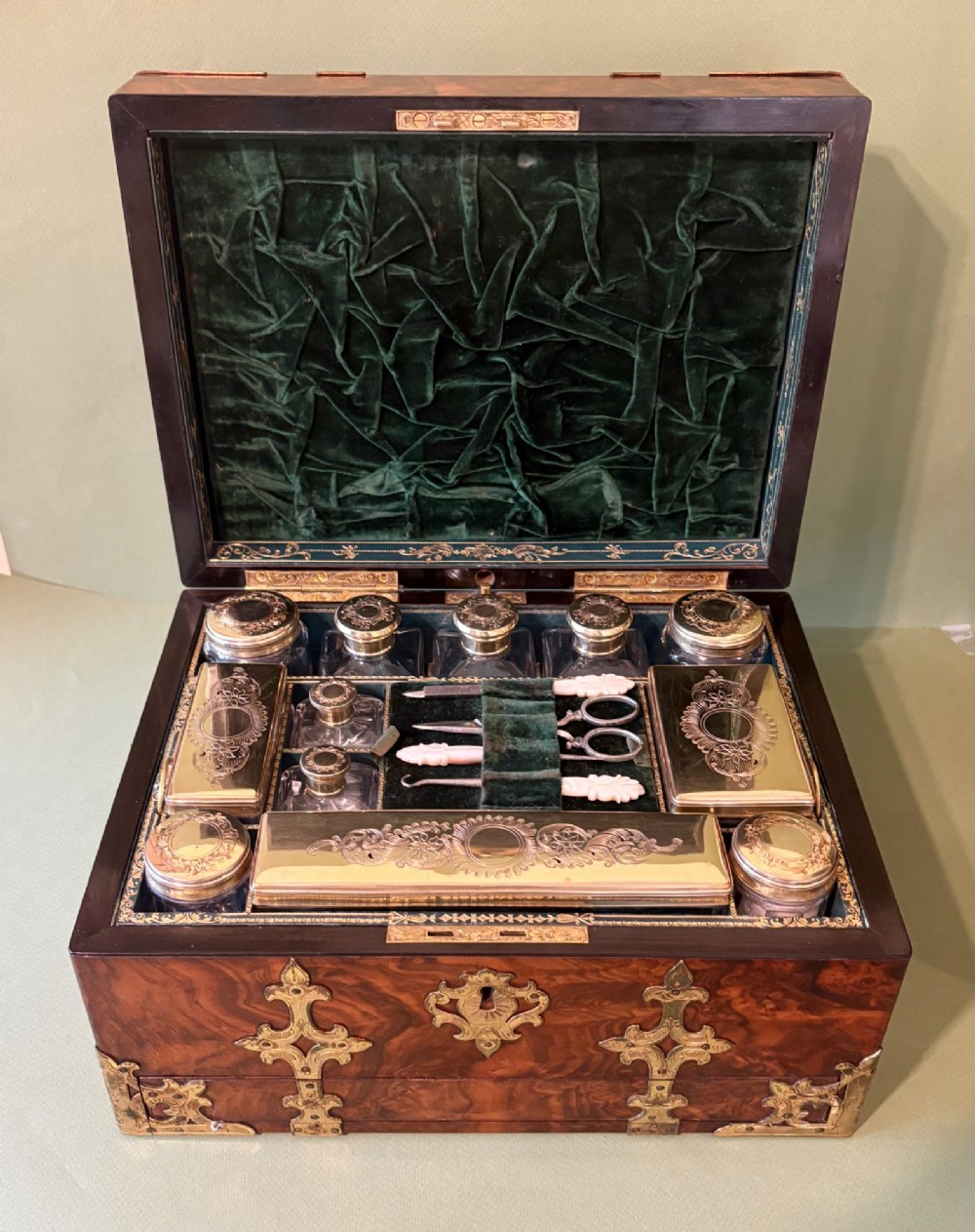 An open Victorian burr walnut dressing case featuring brass mounts, green velvet lining, silver-topped glass jars, vanity tools, and a pull-out lower drawer. View II