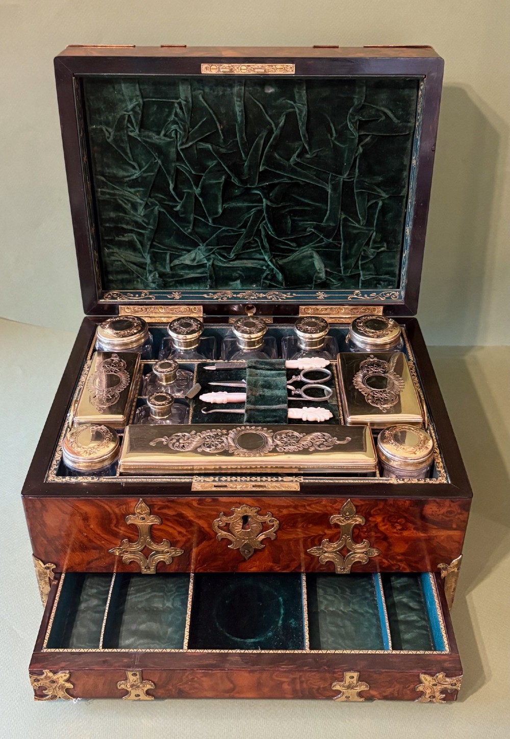 An open Victorian burr walnut dressing case featuring brass mounts, green velvet lining, silver-topped glass jars, vanity tools, and a pull-out lower drawer.