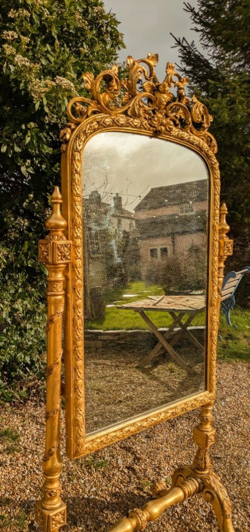 fig 2. An ornate floor-standing cheval mirror framed in gold giltwood with carved cresting and turned supports.