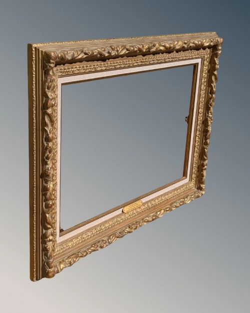 fig 1. A rectangular carved gilt wood picture frame with an inner neutral linen slip and a central brass plaque along the lower edge.