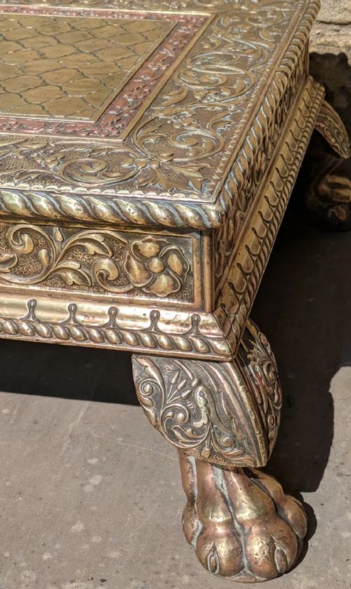 A square antique Indian low table with embossed brass and copper metal cladding, floral foliage motifs, a central geometric panel, and four claw feet. View V