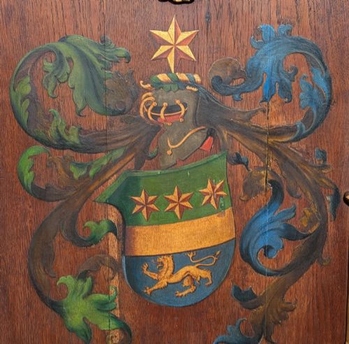 fig 1. A rectangular carved oak wall plaque featuring a hand-painted heraldic coat of arms, gilt scroll moulding, and a Latin motto.