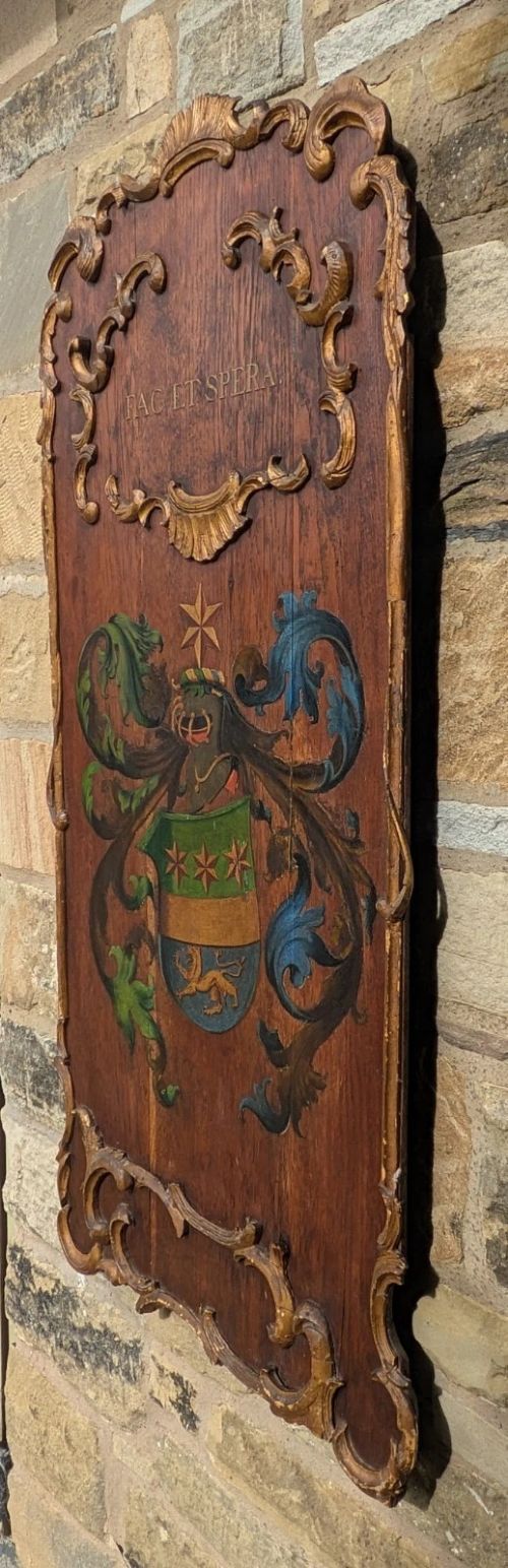 A rectangular carved oak wall plaque featuring a hand-painted heraldic coat of arms, gilt scroll moulding, and a Latin motto. View V