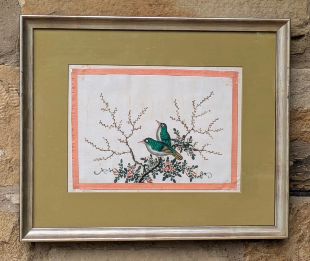 A framed Chinese watercolour painting depicting two green birds perched on flowering branches with an ochre mount and silvered wood frame.