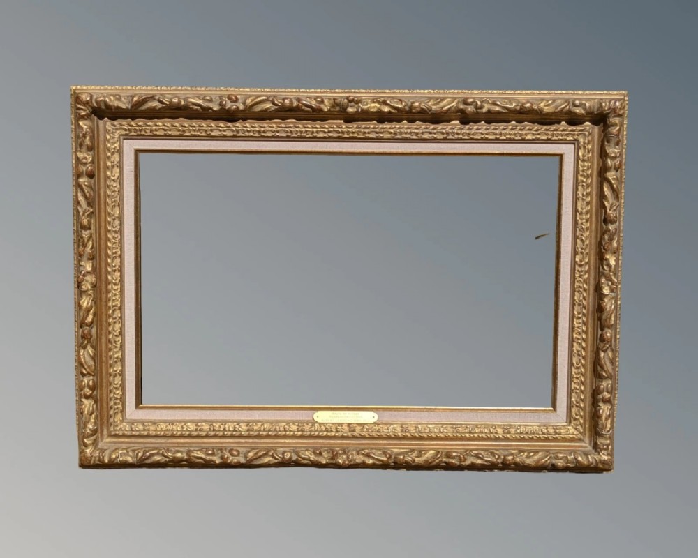 A rectangular carved gilt wood picture frame with an inner neutral linen slip and a central brass plaque along the lower edge.