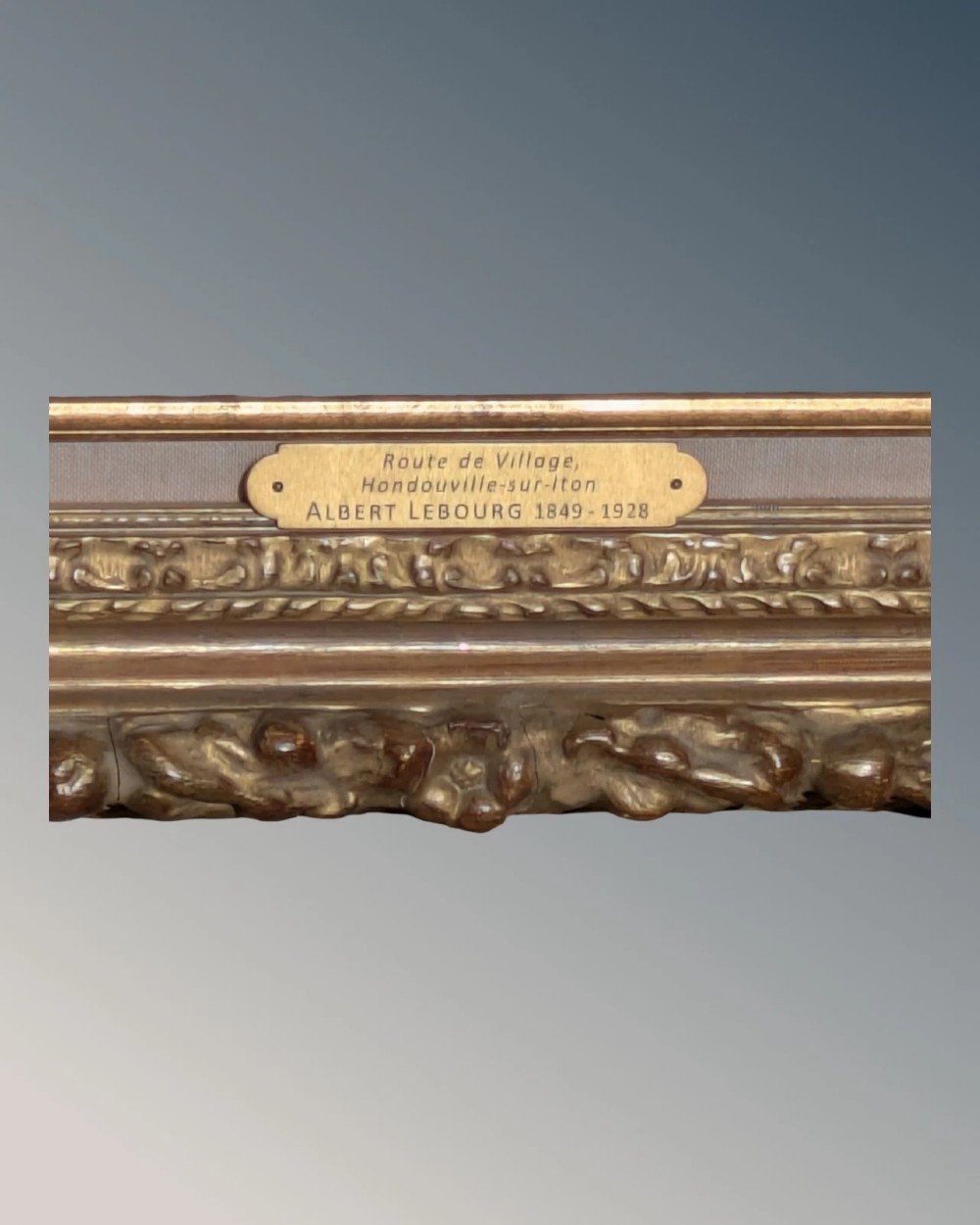 A rectangular carved gilt wood picture frame with an inner neutral linen slip and a central brass plaque along the lower edge. View VI