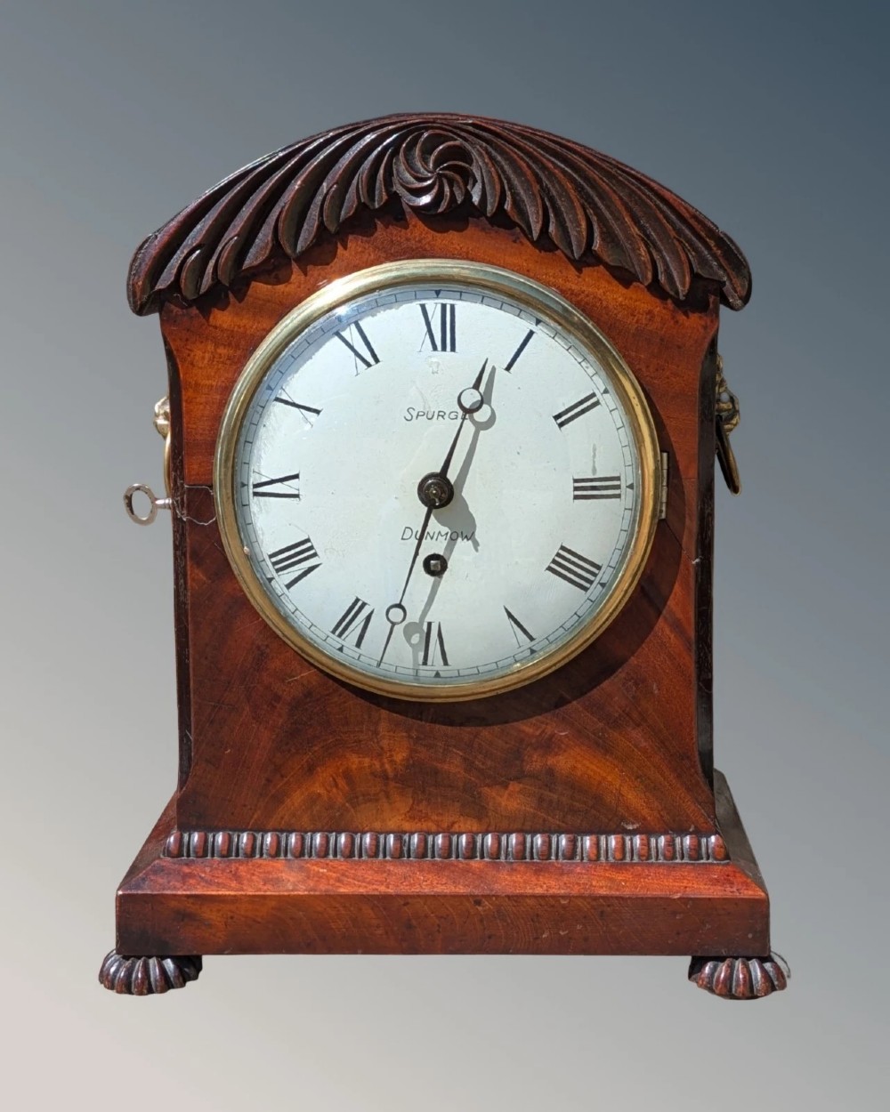 A 19th-century mahogany bracket clock featuring a carved curved pediment, a white circular dial with Roman numerals, side ring handles, and carved bracket feet.