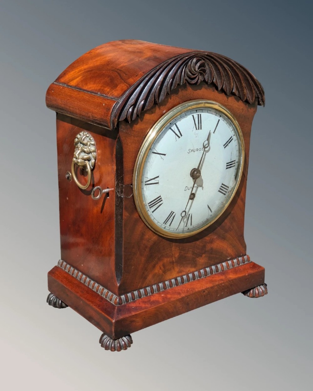 A 19th-century mahogany bracket clock featuring a carved curved pediment, a white circular dial with Roman numerals, side ring handles, and carved bracket feet. View II