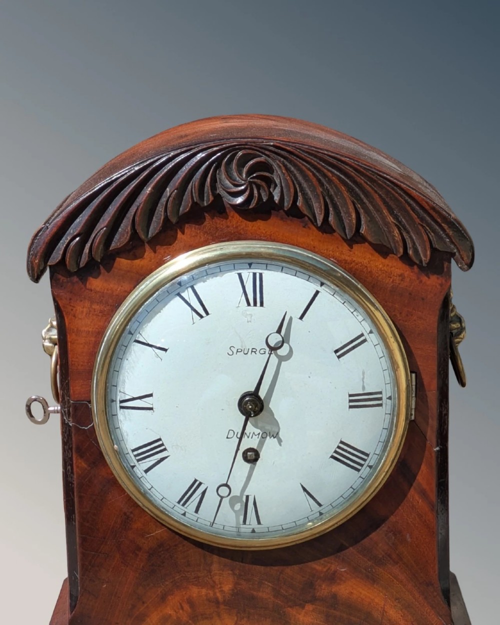 A 19th-century mahogany bracket clock featuring a carved curved pediment, a white circular dial with Roman numerals, side ring handles, and carved bracket feet. View III