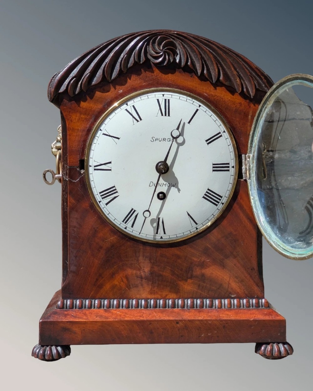 A 19th-century mahogany bracket clock featuring a carved curved pediment, a white circular dial with Roman numerals, side ring handles, and carved bracket feet. View IV