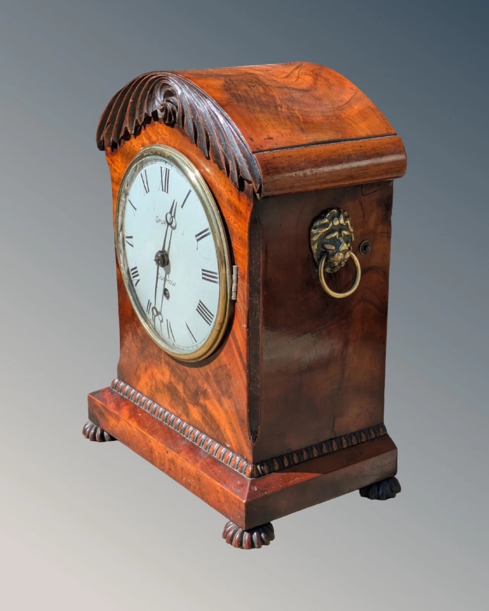 A 19th-century mahogany bracket clock featuring a carved curved pediment, a white circular dial with Roman numerals, side ring handles, and carved bracket feet. View V
