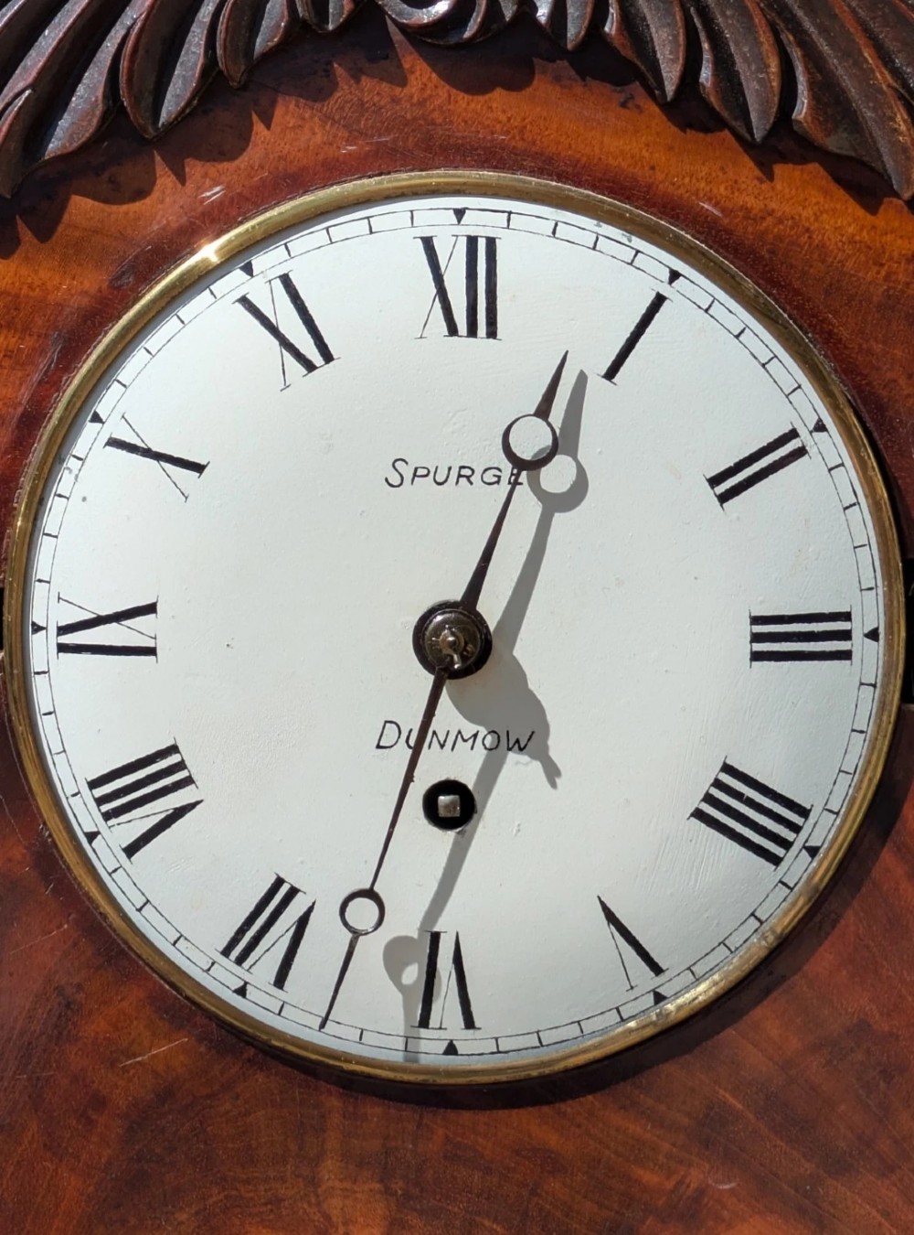 A 19th-century mahogany bracket clock featuring a carved curved pediment, a white circular dial with Roman numerals, side ring handles, and carved bracket feet. View VI