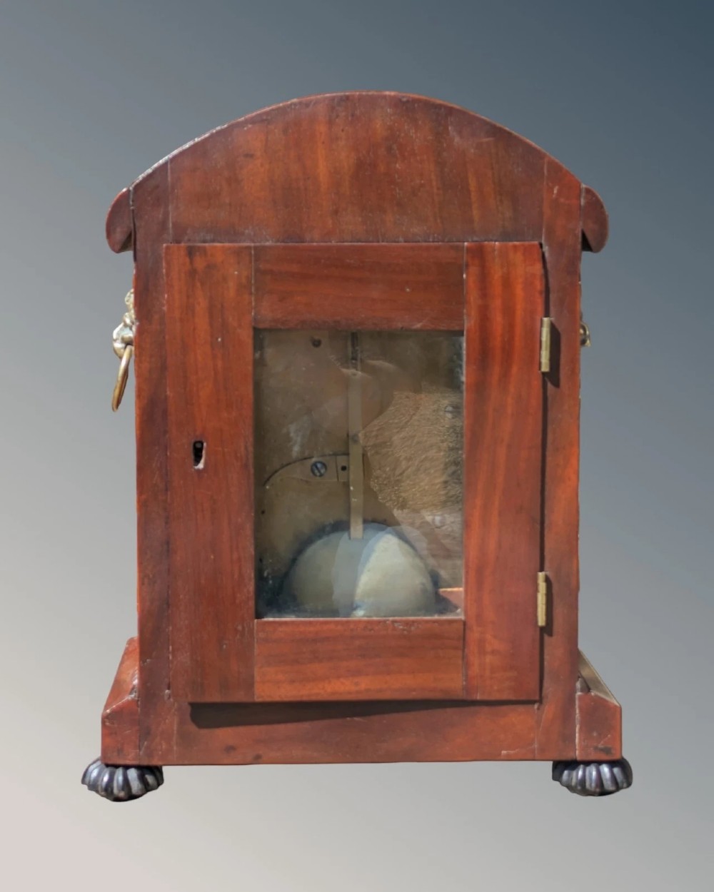A 19th-century mahogany bracket clock featuring a carved curved pediment, a white circular dial with Roman numerals, side ring handles, and carved bracket feet. View VII