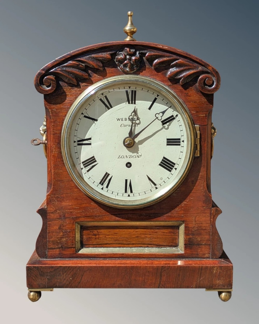 A 19th-century mahogany fusee bracket clock with carved foliate cresting, a brass finial, a white circular dial with Roman numerals, and small brass ball feet.