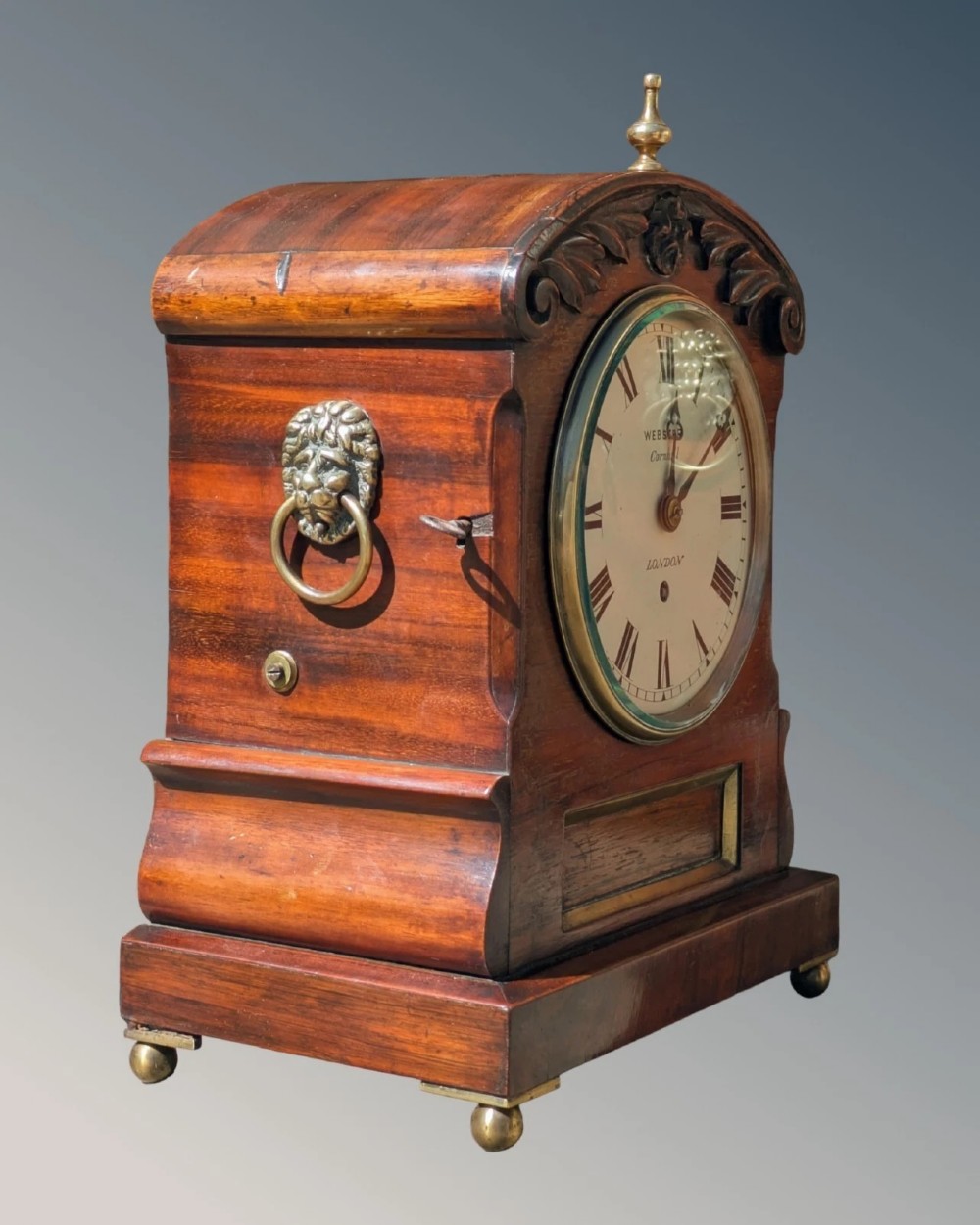 A 19th-century mahogany fusee bracket clock with carved foliate cresting, a brass finial, a white circular dial with Roman numerals, and small brass ball feet. View II