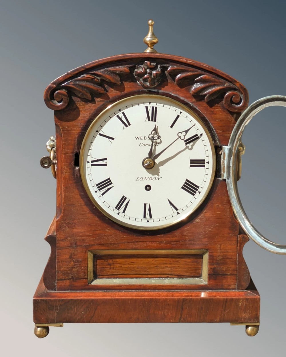 A 19th-century mahogany fusee bracket clock with carved foliate cresting, a brass finial, a white circular dial with Roman numerals, and small brass ball feet. View IV