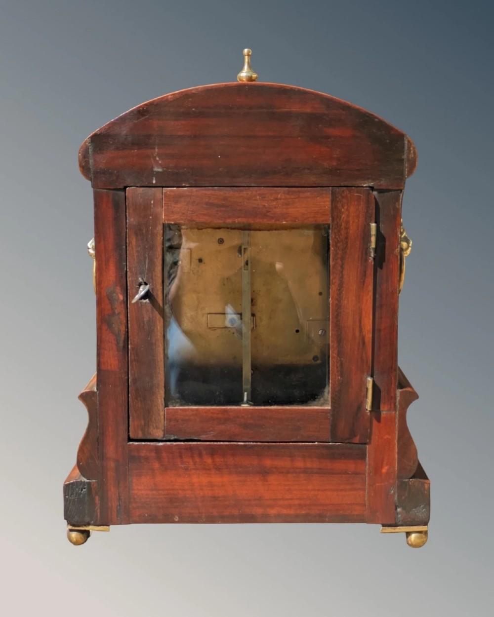 A 19th-century mahogany fusee bracket clock with carved foliate cresting, a brass finial, a white circular dial with Roman numerals, and small brass ball feet. View V