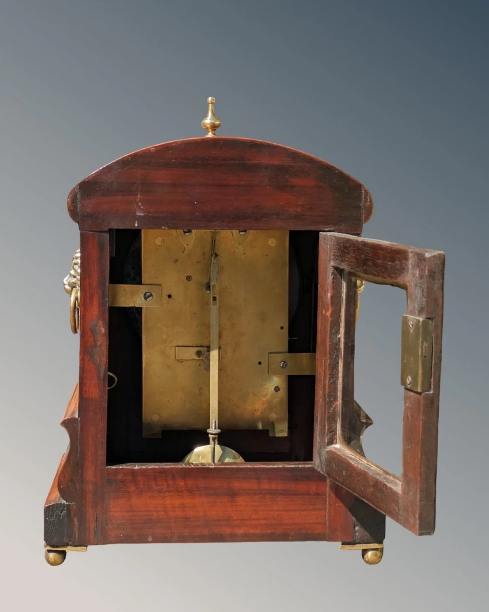 A 19th-century mahogany fusee bracket clock with carved foliate cresting, a brass finial, a white circular dial with Roman numerals, and small brass ball feet. View VI
