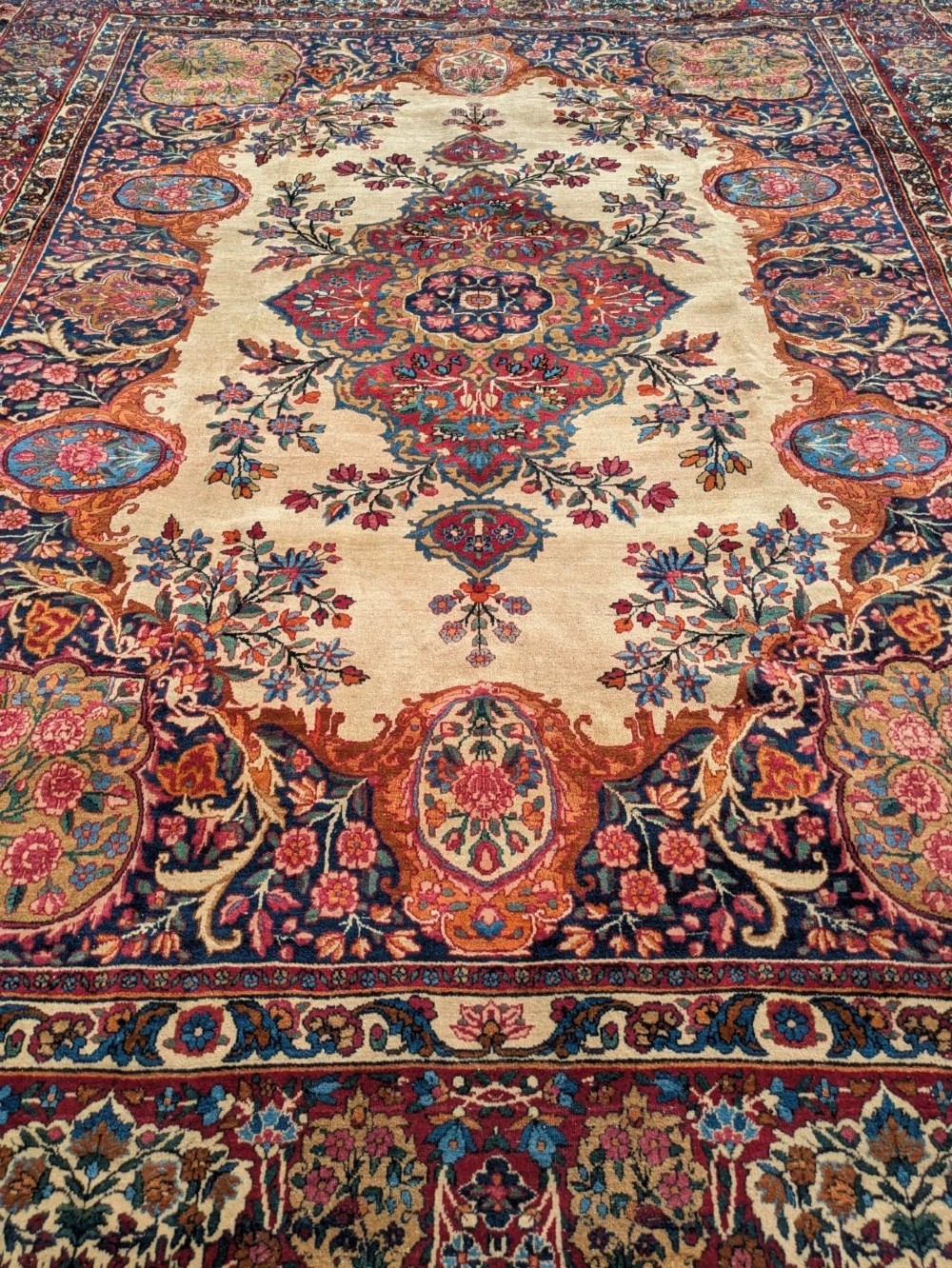 An antique room-sized Kirman Persian carpet featuring a complex central floral medallion on an ivory field surrounded by floral borders in red, navy blue, and gold. View II