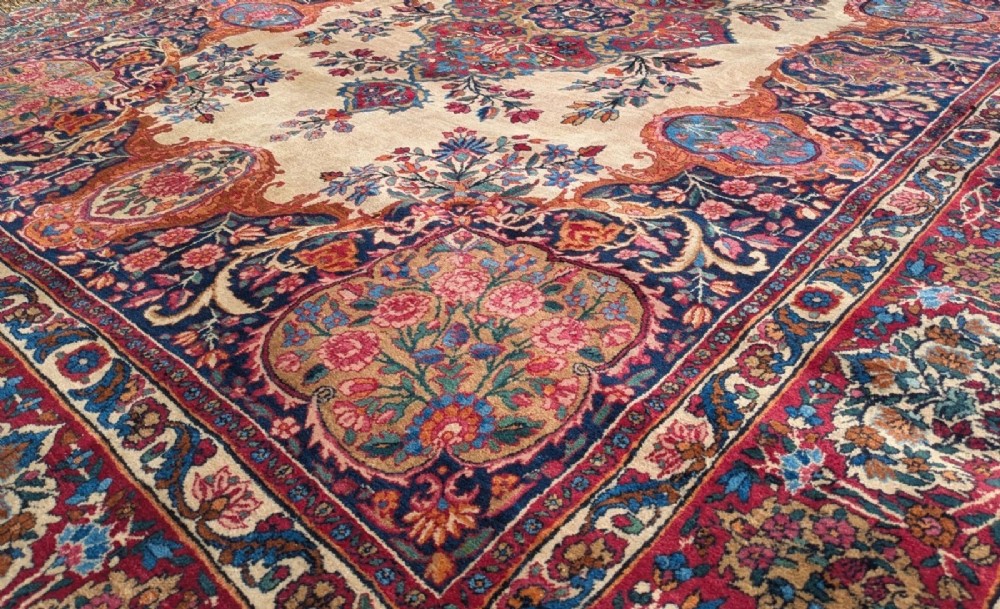 An antique room-sized Kirman Persian carpet featuring a complex central floral medallion on an ivory field surrounded by floral borders in red, navy blue, and gold. View III