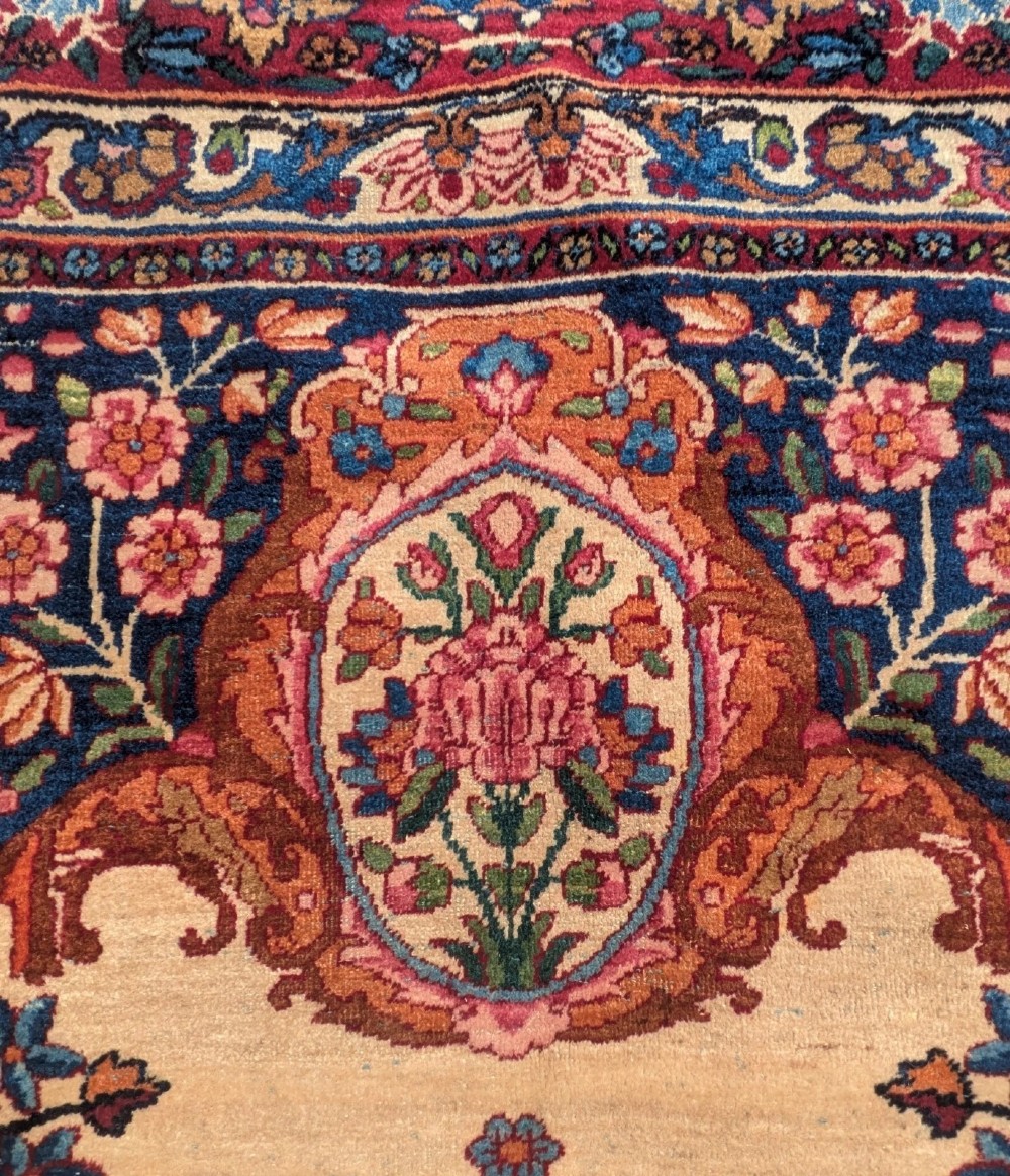 An antique room-sized Kirman Persian carpet featuring a complex central floral medallion on an ivory field surrounded by floral borders in red, navy blue, and gold. View V