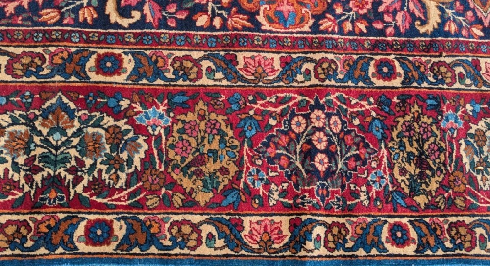 An antique room-sized Kirman Persian carpet featuring a complex central floral medallion on an ivory field surrounded by floral borders in red, navy blue, and gold. View VI