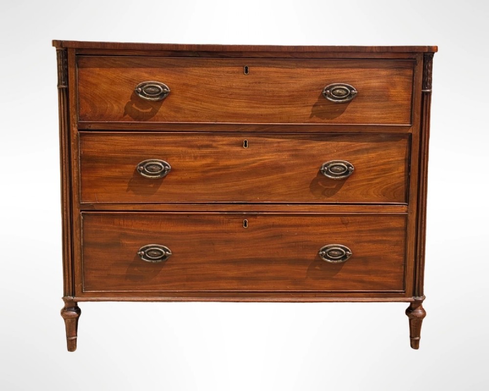 A George III mahogany chest of drawers featuring three full-width drawers with oval brass drop handles, reeded side columns, and turned feet. View II