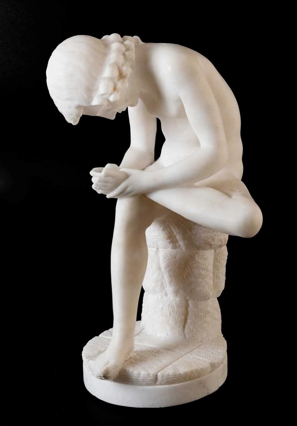 A carved white alabaster sculpture depicting a classical nude youth seated on a tree stump while extracting a thorn from his foot.