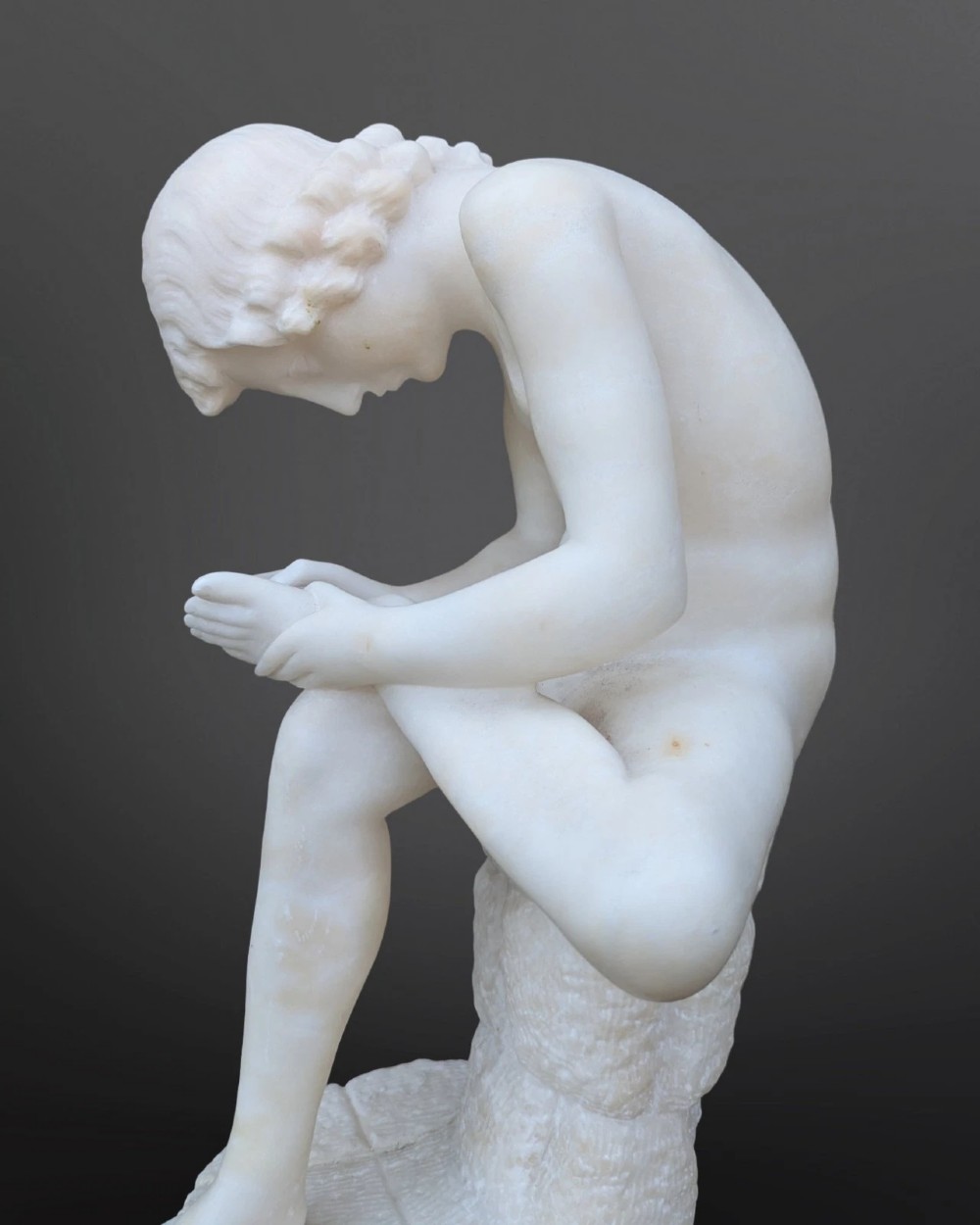 A carved white alabaster sculpture depicting a classical nude youth seated on a tree stump while extracting a thorn from his foot. View IV