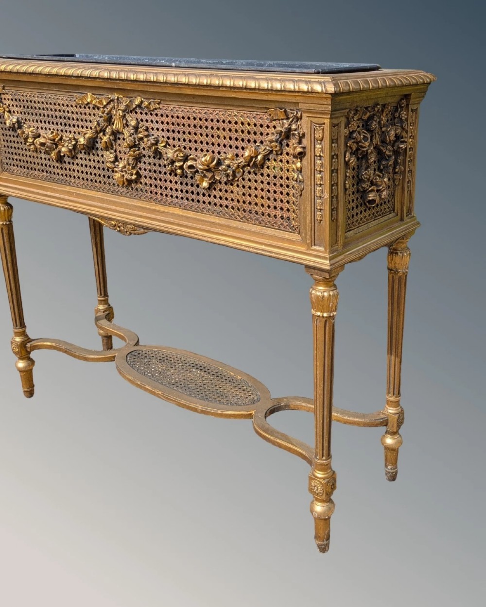 An antique Louis XVI style giltwood and cane jardiniere stand featuring carved floral swags, fluted legs, and an oval stretcher. View VI