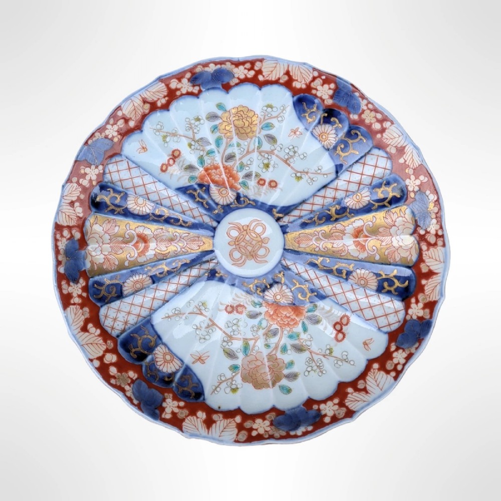 A circular Japanese Imari porcelain plate featuring a scalloped rim, fluted surface, and floral motifs in cobalt blue, iron red, and gold decoration.