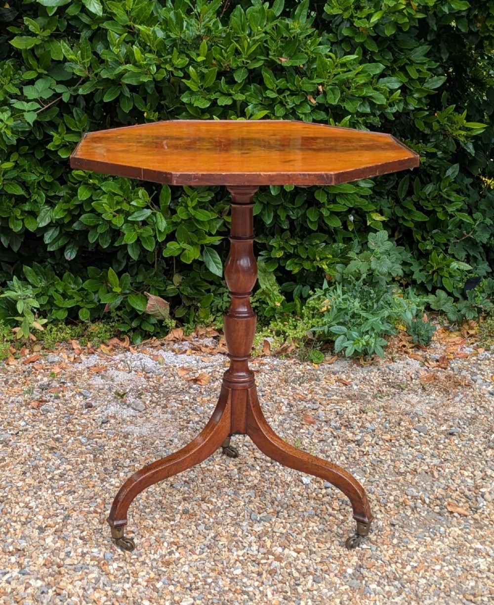 An antique Regency mahogany occasional table featuring an octagonal tilt-top surface, a turned central pedestal stem, three splayed tripod legs, and brass castors.